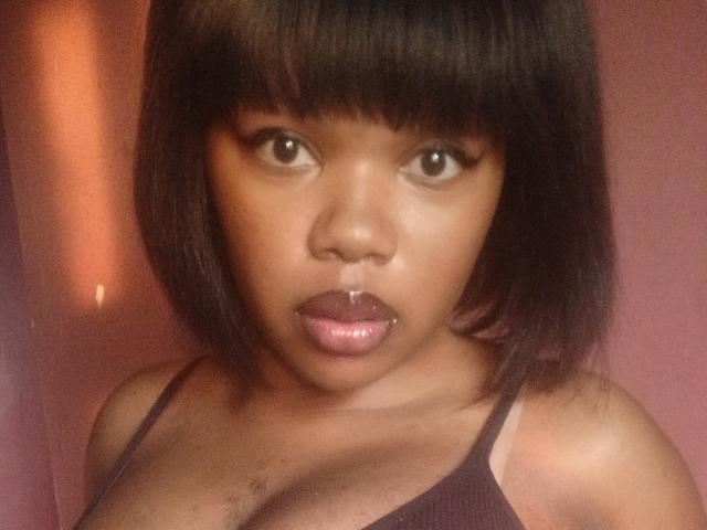 KatyBTeresa has a dark brown bob with bangs, a nose ring, and shiny pink lips. She's wearing a black tank top, showing her small tits and a bit of chest hair.