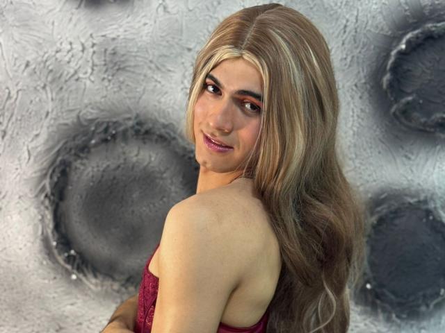 MichellPratTrans with long blonde hair, red lipstick, and red lace top, looking back with a slight smile, showing bare shoulders and a cute face.