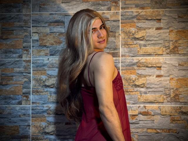 MichellPratTrans with long blonde hair, red lace top, looking over shoulder, smiling at camera, standing against stone wall, showcasing slim arms and subtle makeup.