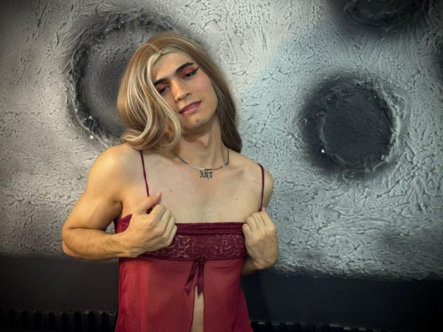 MichellPratTrans with blonde wig, red eyeliner, and maroon lingerie, pulling up the top to expose their small, flat tits. Standing, hands on chest, wearing a silver necklace.