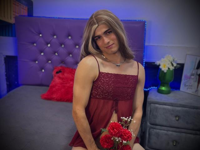 MichellPratTrans with long blonde hair, wearing a red lace babydoll, holding red roses, sitting on a bed with a purple tufted headboard, and a gray nightstand with flowers.