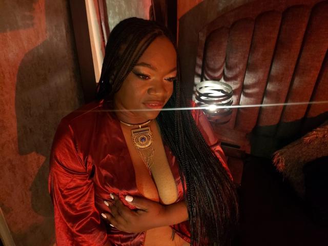 RoxiCoquette in a red velvet robe, holding her large, dark brown tits, with long braided hair, wearing a gold necklace, in a dimly lit, sexy, intimate setting.