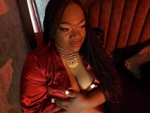 RoxiCoquette in a red satin robe, showing her big tits with her hands, wearing a gold necklace with a blue emblem, looking down with a slight smile, long braids.