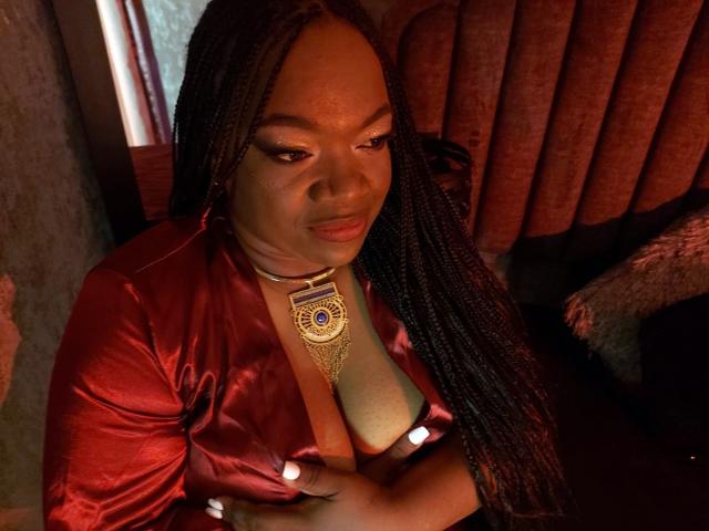 RoxiCoquette with dark skin, long braids, wearing a red satin shirt, exposing her tits with a gold necklace, looking down with a slight smile in dim light.