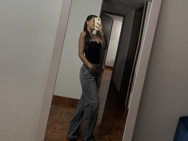 ThaliaAres with long black hair, wearing a black tank top and gray flared jeans, taking a mirror selfie, showing a slim figure and small tits.
