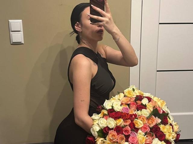NikkiGoldX with dark hair in a black dress, holding a large bouquet of roses, taking a mirror selfie, showing her tits and ass, in a room.