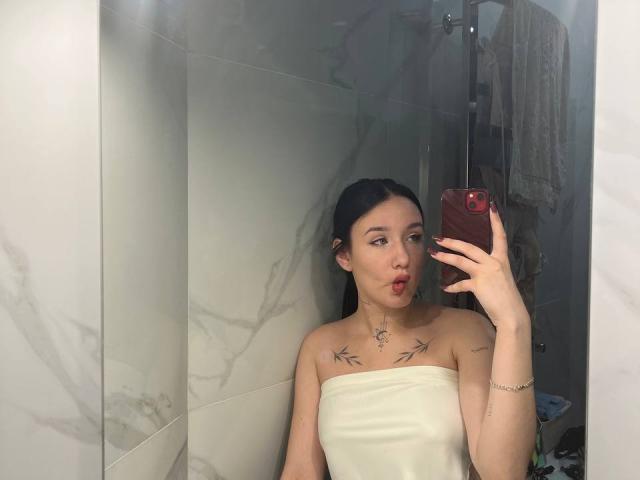 NikkiGoldX with dark hair, pouty red lips, white strapless top, bird tattoos on chest, holding red phone, mirror selfie, marble walls, small silver bracelet on right wrist.