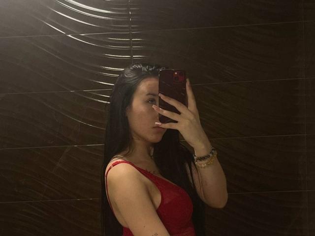 NikkiGoldX, con cabello negro largo, vistiendo una camiseta sin mangas roja, sosteniendo un teléfono frente a su cara, mostrando sus pechos en un selfie frente al espejo, pared de azulejos oscuros..