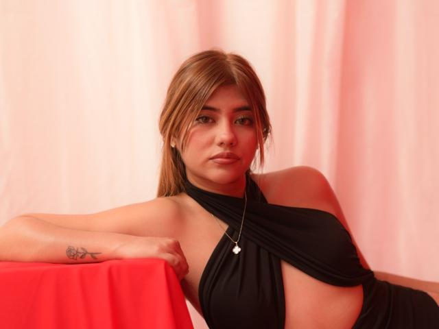 ElySainz with brown hair, black halter top, small tattoos on left arm, leaning on red cloth, wearing silver necklace, looking at camera, soft pink background.