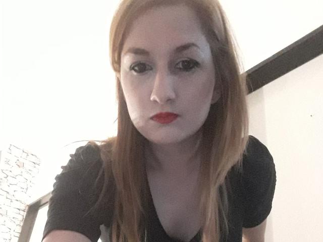 LuzyHot with pale skin, red lipstick, and long brown hair, looking down at the camera with a serious expression, wearing a black shirt, in a slightly blurred photo.