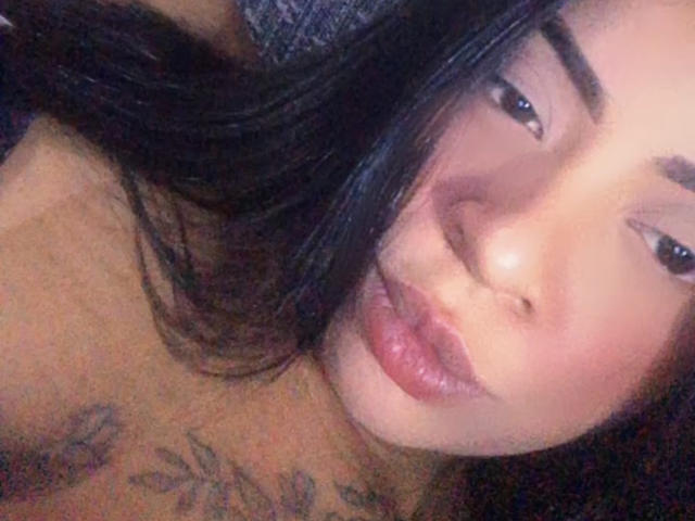 KimberlyAngel's pouty face with glossy lips, dark eyebrows, and long black hair covering part of her face. Tattoo visible on her chest. Her brown eyes are slightly squinted.