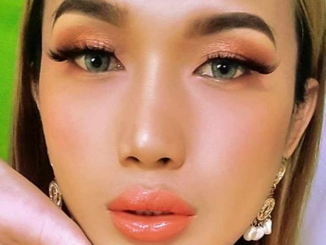 xGoddessJazzyx heeft glanzende oranje lippen, groene ogen met lange wimpers en bruine wenkbrauwen. Ze draagt sierlijke oorbellen en heeft een gladde, glanzende huid. Haar hand is dichtbij haar gezicht..