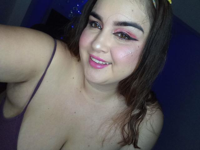 PattyHot69 with pink eyeshadow, dark eyebrows, and pink lipstick, smiling at the camera, showing her big tits and cleavage in a purple top. Her brown hair is down.