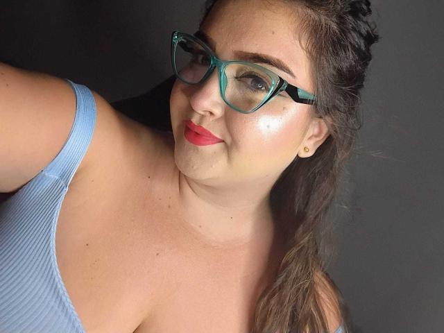 PattyHot69 with glasses, red lipstick, and blue ribbed top, showing her tits, light skin, braided brown hair, and subtle makeup, smiling slightly, against a dark background.