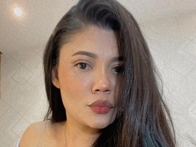 Maryorieth has long brown hair, dark eyebrows, and pink lips. She's looking at the camera with a neutral expression, wearing a white top, and has a light skin tone.