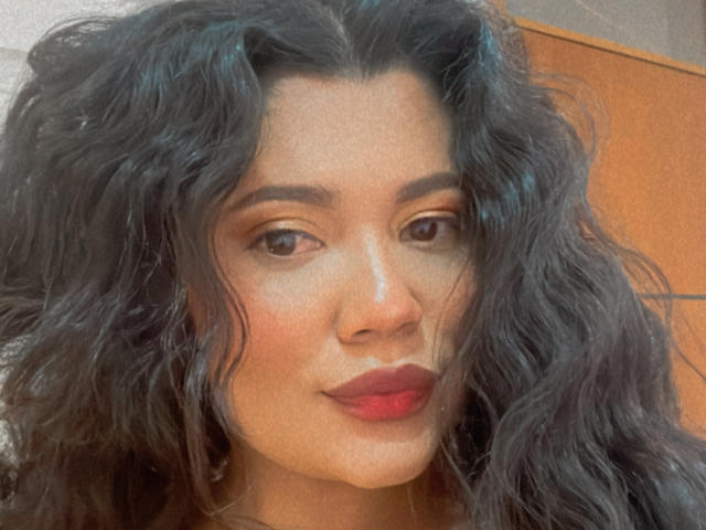 Maryorieth has wavy black hair, brown eyes, and red lipstick. Her skin is tan, and she's smiling subtly. Her nose is small and cute. The photo is slightly grainy.