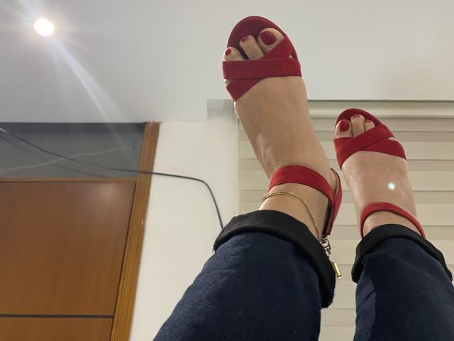 Maryorieth's legs in red open-toe heels with ankle straps, wearing dark blue jeans, showing red-painted toes, and a gold ankle bracelet. Ceiling light and door in background.