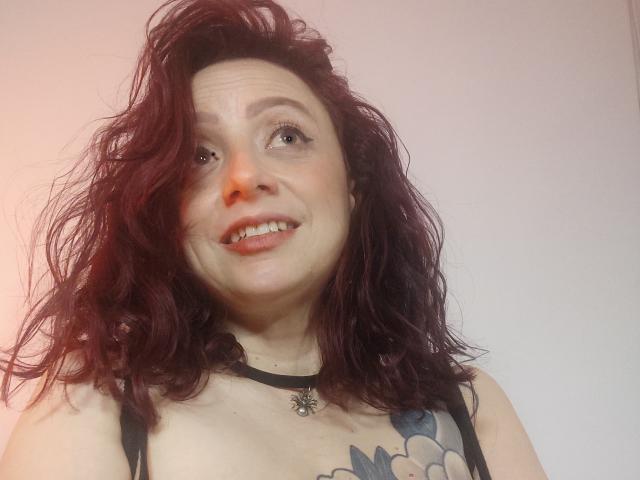 CatalinaMonyque has messy red hair, smiling with freckles, wearing a black choker with a flower, and a tattoo on her chest showing large white flowers.