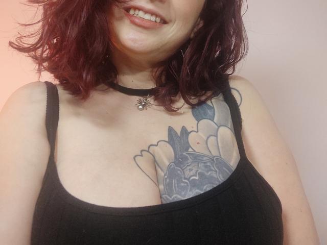 CatalinaMonyque with red, wavy hair, smiling, wearing a black tank top, showing a large, colorful tattoo of hands holding a dark, abstract face on her tits.