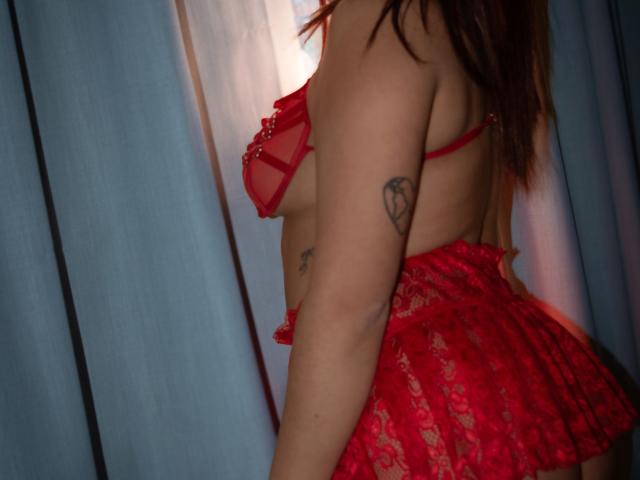 LadyStarOne trägt rote Spitzendessous, zeigt ihre Brüste und ihren Hintern, hat ein Herz-Tattoo auf ihrem rechten Arm, ihr braunes Haar ist offen, und sie befindet sich in einem schwach beleuchteten Raum..