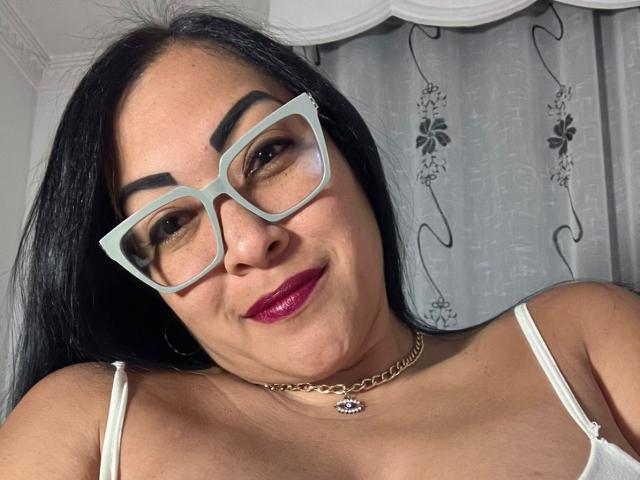 MellissaCortez has dark hair, wears green glasses, and red lipstick, with white straps on her chest, a gold chain necklace, and a small pendant, smiling at the camera.