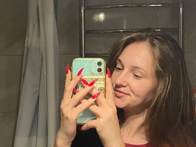 PamelaXu with long brown hair, red nails, and a cute smile, holding a phone with a green case, taking a mirror selfie, showing off her tits.