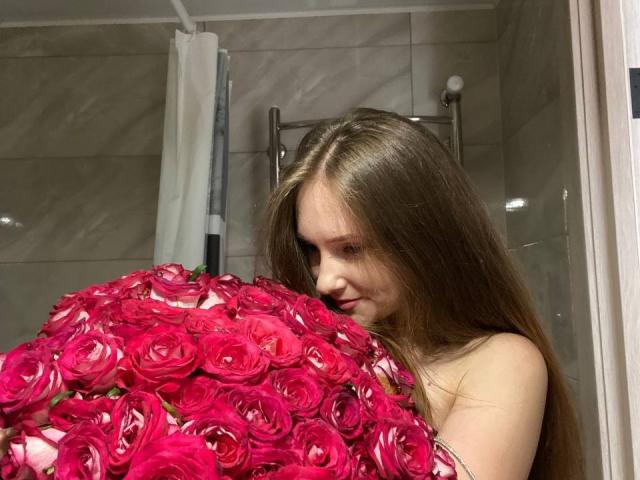PamelaXu with long brown hair holding a massive bouquet of pink roses, covering her bare tits with the flowers. She's in a bathroom with grey tiles.