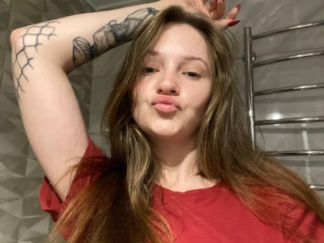 PamelaXu with long brown hair, red shirt, pouty lips, raising right arm showing tattoo, gray wall, and chrome ladder in background. Her tits are small, red nails.
