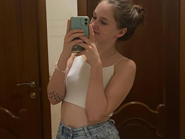 PamelaXu with small tits in a white crop top, holding a phone, wearing denim shorts, standing in front of a mirror, brown hair in a bun, wooden door background.