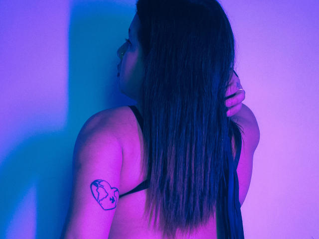 LadyStarOne with long brown hair, back to camera, black tank top, heart tattoo on upper left arm, purple-blue lighting, hand in hair, looking to the left.