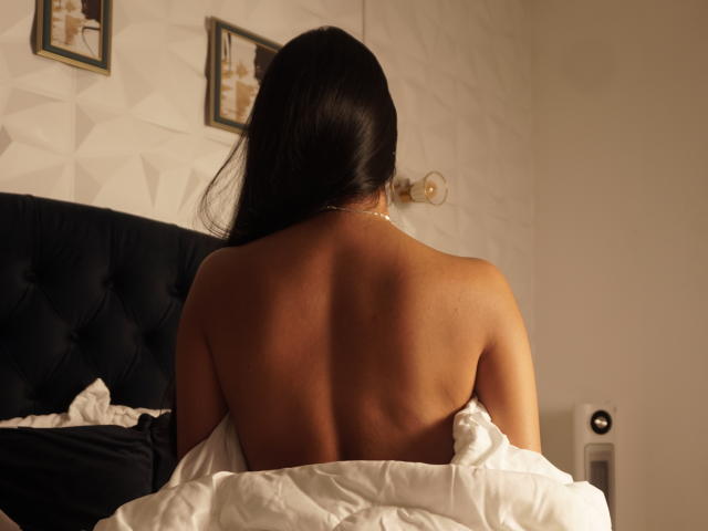 SamanthaRosses, back to the camera, bare ass, tan skin, long black hair, white bedsheet, dark blue headboard, gold wall sconce, framed pictures, subtle lighting, cozy bedroom.