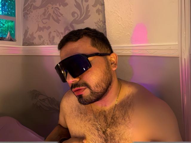 GaelB mit einer behaarten Brust, der schwarze Sonnenbrillen und eine goldene Halskette trägt, blickt nach unten und hat den Mund leicht geöffnet. Warme, violette und rosa Lichter beleuchten sein Gesicht..