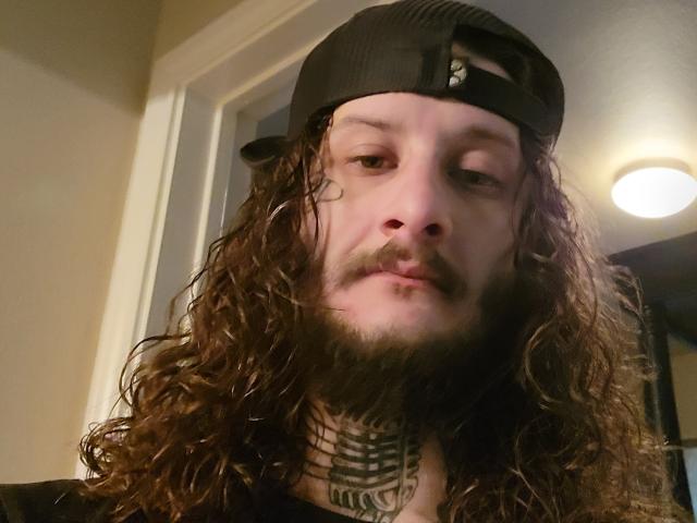 Devon69X with long, curly brown hair, black baseball cap, beard, tattoos on neck, slightly droopy eyes, and a neutral expression, wearing a black shirt.