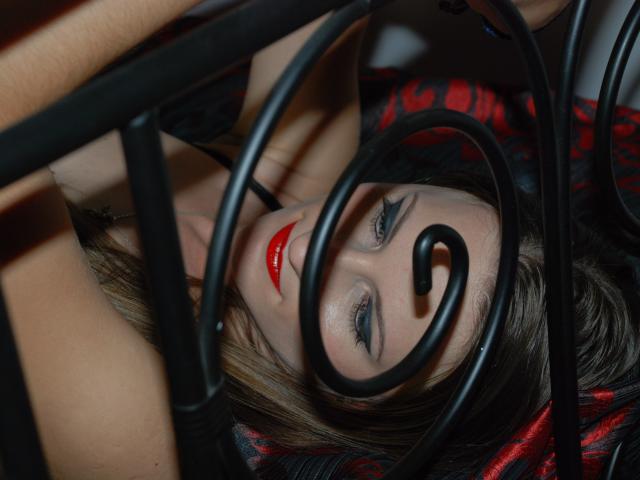 Il volto di AbyX è parzialmente incorniciato da barre di metallo nero, con labbra rosse, occhi azzurri e capelli castano scuro, e giace su una superficie con un motivo rosso e nero..
