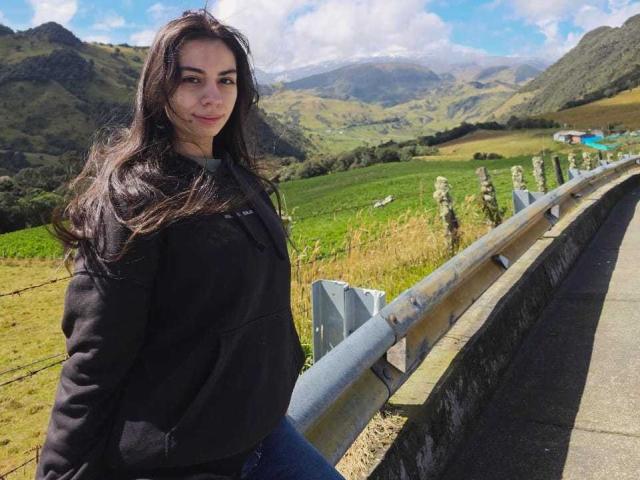 KatherinJohns stands by a metal fence, wearing a black hoodie, with long dark hair, and a slight smile, in a sunny, green, mountainous landscape. Her tits are small.