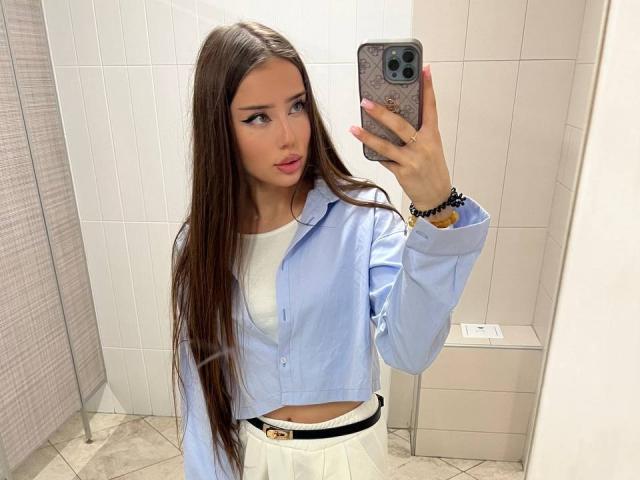 ImmyMegan, avec de longs cheveux bruns, une chemise bleue, un haut blanc, une jupe blanche, tenant un téléphone, selfie devant un miroir, regardant la caméra, lèvres roses, ceinture noire, bracelet, salle de bain carrelée, expression sérieuse..