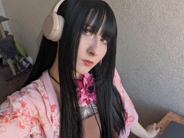 MelissaRaven with long black hair, pink floral kimono, beige headphones, green eyes, pink flower, pale skin, holding tablet, cat tree in background.