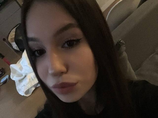 Amabel has long brown hair, dark eyebrows, and is pouting with slightly parted lips, wearing black eyeliner. Her nose is pierced on the right side. Her skin is light.