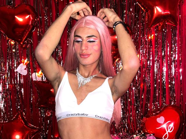NohemiWon with pink hair, white bra, and sparkling necklace, arms raised, showing armpits, wearing pink eyeshadow and lipstick, smiling, surrounded by red heart balloons.