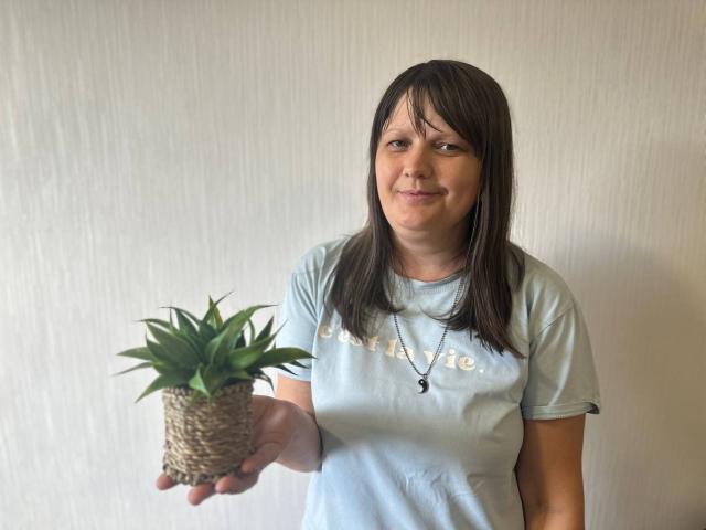 Susan Bim sosteniendo una pequeña planta verde en una cesta de mimbre, vistiendo una camisa azul claro con un collar, sonriendo ligeramente, cabello castaño, piel clara, de pie frente a una pared lisa..