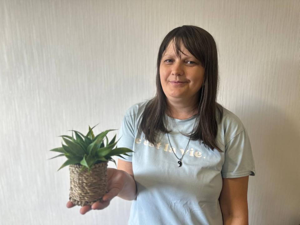 SusanBim holding a small green plant in a woven basket, wearing a light blue shirt with a necklace, smiling slightly, brown hair, light skin, standing in front of a plain wall.