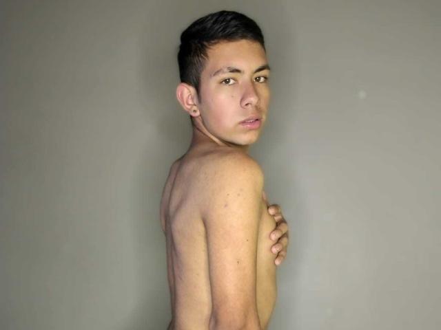 AdamMatt, sem camisa, com cabelo preto curto, olhos castanhos e pele clara, cobrindo seus pequenos seios nus com a mão direita, olhando por cima do ombro, em um quarto cinza neutro..