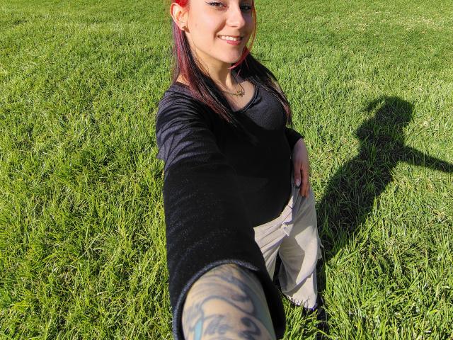 AmyStreetwise69 with red hair, smiling, black top, beige pants, tattooed arm, reaching out for selfie, shadow on grass, sunlight, green grass, pregnant belly, smiling, outdoor.