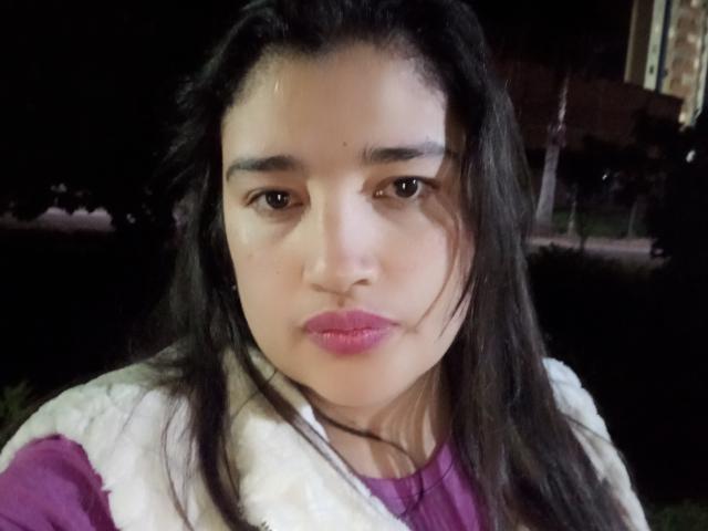 A ZafiRouge tem olhos castanho-escuros, batom rosa e cabelos longos e pretos. Ela está vestindo uma camisa roxa com um colete branco e fofo. O rosto dela está levemente suado..