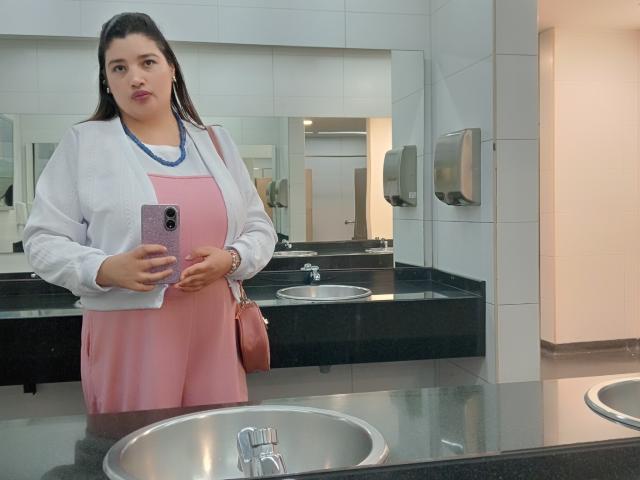 ZafiRouge se tient dans une salle de bain, en train de prendre un selfie. Elle porte une robe rose, un cardigan blanc et un sac à main rose. Elle a une poitrine de taille moyenne, des cheveux bruns et une expression sérieuse..