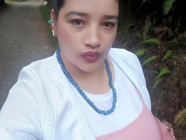 ZafiRouge a une poitrine de taille moyenne, elle porte un tablier rose par-dessus une chemise blanche, un collier de perles bleues et des boucles d'oreilles vertes, avec des cheveux foncés et du rouge à lèvres rose, sur une photo d'elle-même..