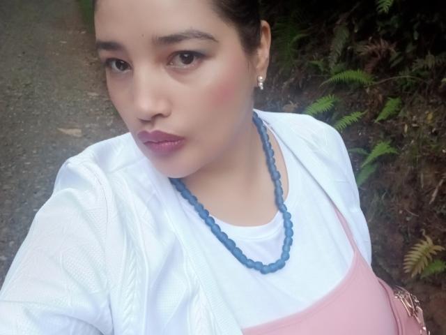 ZafiRouge has light skin, dark hair, and brown eyes. She's wearing a pink apron over a white shirt, blue bead necklace, and pink lipstick. Her tits are small.