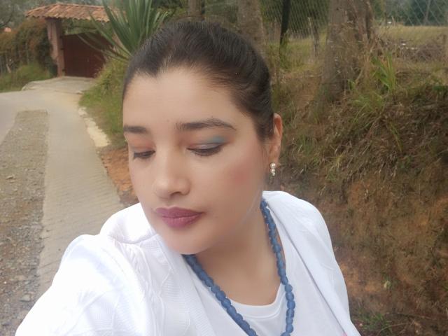 ZafiRouge with dark hair, blue eyeshadow, pink lipstick, white shirt, blue bead necklace, and earrings, looking down with closed eyes. Her tits are small, and she's cute.