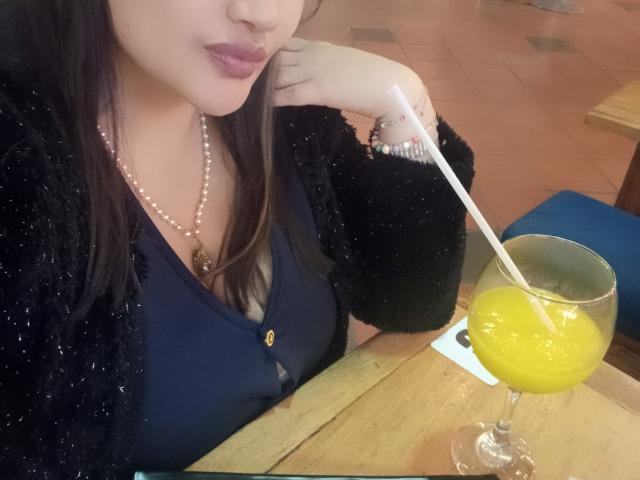 ZafiRouge with her dark hair, black sparkly cardigan, and pearl necklace, sipping a yellow cocktail with a white straw, her plump pink lips slightly parted.