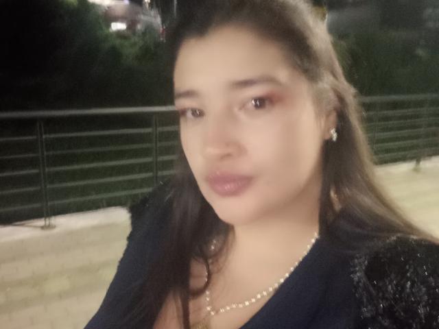 ZafiRouge has medium brown skin, dark brown hair, pink eyeshadow, glossy pink lips, wearing a black dress with sparkles, pearl necklace, and diamond earrings, in a blurry photo.